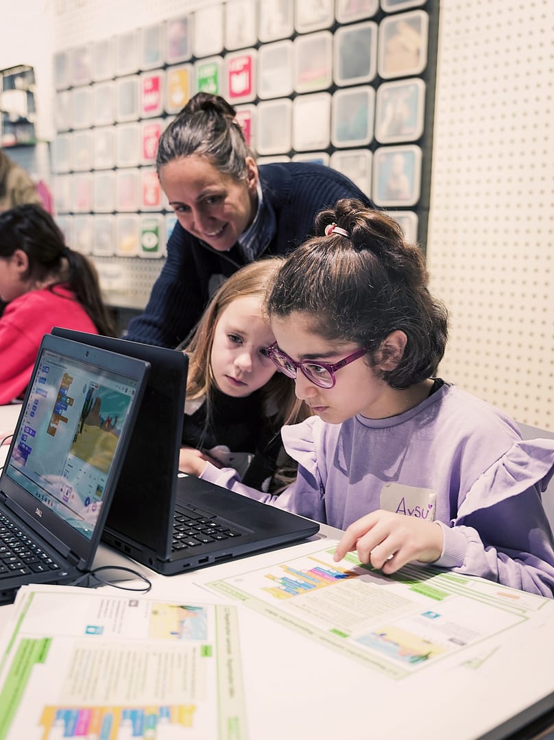 Zwei Mädchen, die an einem Tisch sitzen und zusammen auf einen Laptop schauen. Hinter ihnen eine Frau, die den Mädchen über die Schulter schaut.