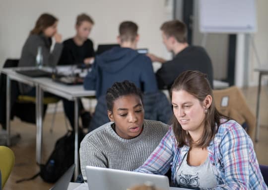 Mädchen vorne an einem Rechner im Hintergrund Gruppe Jugendlicher